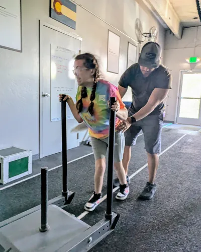 A trainer assists a client pushing a weighted sled during gait training