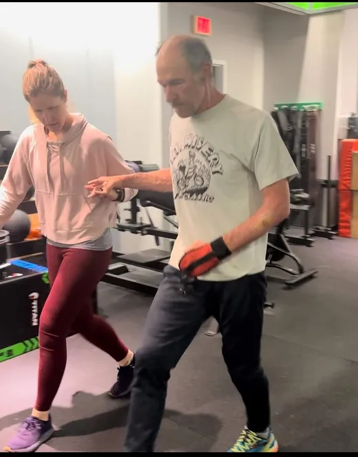Trainer Liz walks side by side with an athlete, holding his hand for support during gait and balance training