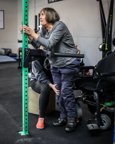A trainer helps a client practice standing balance at a support rack, with a wheelchair nearby